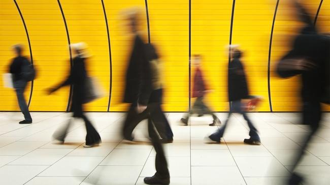 Commuters walk through a subway station tunnel with bold yellow walls.