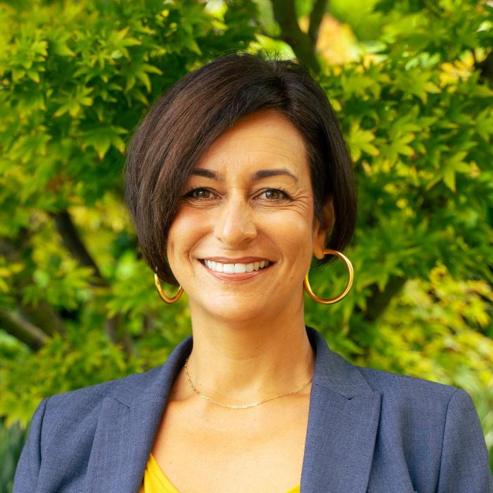 Kristina's headshot, she is a Latina woman with a short angled bob haircut and a warm smile. She wears gold hoop earrings and a blue suit jacket